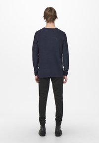 Homme debout de dos, portant une chemise à manches longues bleu marine, un pantalon noir et des bottes noires sur un fond blanc uni.
