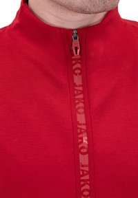 Haut de sport zippé rouge en tissu lisse, avec un col haut, une fermeture éclair contrastante et "Jako" imprimé verticalement le long de la fermeture éclair.