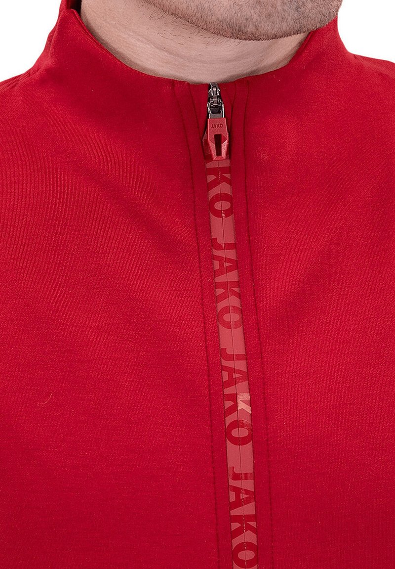 Haut de sport zippé rouge en tissu lisse, avec un col haut, une fermeture éclair contrastante et "Jako" imprimé verticalement le long de la fermeture éclair.