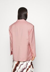 Veste rose clair avec un design structuré, col cranté et manches longues. Texture lisse avec des fentes latérales à l'ourlet pour faciliter les mouvements.