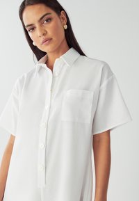 Bela majica s kratkimi rokavi, gumbi na sprednji strani, enim žepom na prsih ter mehko teksturo tkanine. Ima velike gumbe in oblikovan ovratnik.