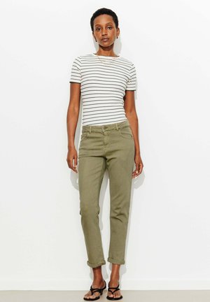 Femme portant un t-shirt rayé blanc et noir, un pantalon olive retroussé et des sandales noires à lanières, debout contre un mur blanc.