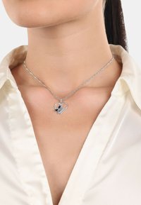 Srebrny naszyjnik z serduszkiem w kształcie pendanta z niebieskimi akcentami na skręcanym łańcuszku, prezentowany na jasnej bluzce.