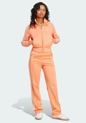 Tuta arancione con accenti e righe bianche, giacca con zip e colletto alto, e pantaloni a sigaretta. Caratterizzato da un tessuto morbido e liscia e tasche.