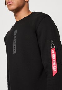 Homme portant un sweat-shirt noir avec le texte vertical "ALPHA" sur la poitrine et une étiquette de fermeture éclair rouge "REMOVE BEFORE FLIGHT" sur la poche de la manche supérieure.