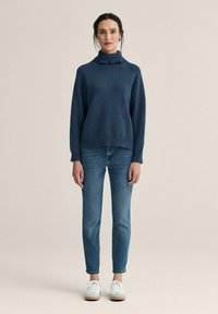 Laivastonsininen neulottu korkeakauluksinen paita, jossa on resorit helmassa ja hihansuissa, yhdistetty kapealahkeisiin sinisiin farkkuihin ja valkoisiin lenkkareihin, joissa on kumipohjat.