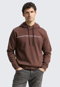Bruine hoodie met trekkoorden, voorzien van witte en rode horizontale strepen over de borst. Soepele textuur, relaxte pasvorm en kangoeroezak.