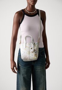 Handtasche mit weißem Blumenmuster, doppelten Griffen und einem verstellbaren Schultergurt. Ausgestattet mit goldfarbenen Beschlägen und einem kleinen Anhänger.