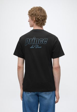 Jeune homme aux cheveux bouclés porte un T-shirt noir avec le texte bleu « prince Les Deux » dans le dos, associé à un jean bleu, debout sur fond blanc.