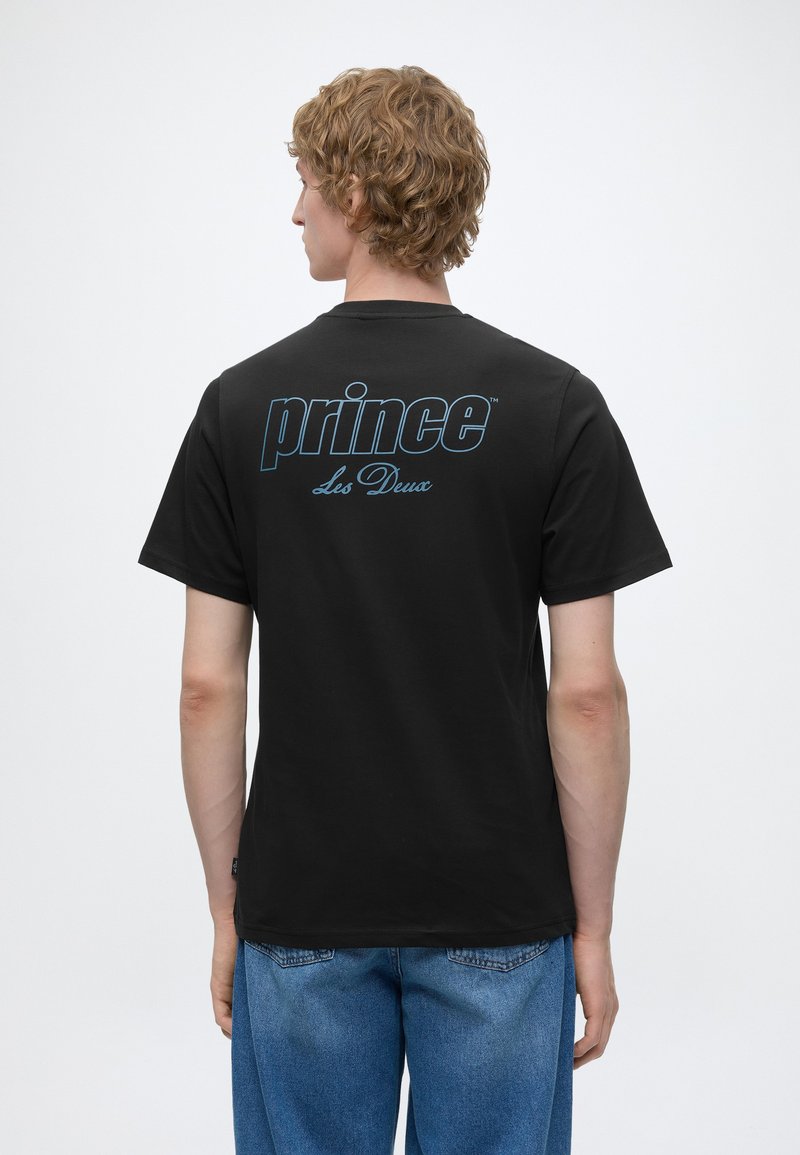 Νέος άνδρας με σγουρά μαλλιά φοράει μαύρο T-shirt με μπλε κείμενο "prince Les Deux" στην πλάτη, σε συνδυασμό με μπλε τζιν, στέκεται μπροστά σε λευκό φόντο.