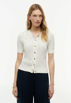 Mujer con cabello largo y rubio, vistiendo un cárdigan blanco de manga corta con botones y pantalones oscuros, de pie frente a un fondo claro y liso.