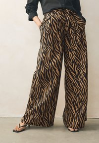 Pantalones de pierna ancha con un audaz estampado de rayas de tigre en negro y caramelo. La tela parece suave con un ligero brillo. Cuenta con bolsillos laterales.