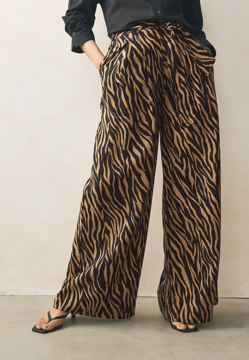 Pantalones de pierna ancha con un audaz estampado de rayas de tigre en negro y caramelo. La tela parece suave con un ligero brillo. Cuenta con bolsillos laterales.