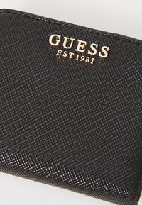 Portefeuille texturé noir avec un logo doré "GUESS EST 1981". Le matériau durable présente un motif subtil et une forme rectangulaire élégante.