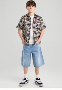 Camisa beige de manga corta con botones y estampado de hojas en azul marino, camiseta blanca, pantalones cortos de mezclilla azul claro y zapatillas negras con detalles blancos.