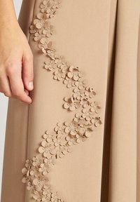 Vestido de tela beige con un diseño de apliques florales a lo largo del costado, que presenta pequeñas flores en capas realizadas en un material ligeramente texturizado.