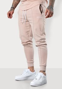 Helle rosa Jogginghosen mit weißen Kordeln, Seitentaschen und gesticktem "ESTEEM"-Logo. Tragen mit weißen Sneakern. Glattes Material, schlanker Schnitt.