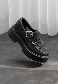 Sapatos de couro preto laqueado com uma faixa com fivela, design frontal trançado, sola de borracha grossa e detalhes em costura branca.