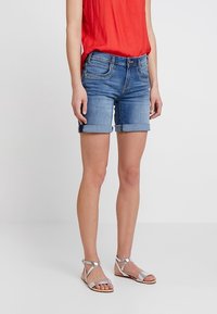 Denim shorts, light blue, rolled hem, five-pocket design, paired with silver strappy sandals. Model wearing a red top.