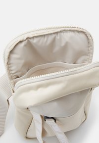 Beige axelväska med öppen dragkedja som visar en innerficka och justerbar rem med tryckt text.