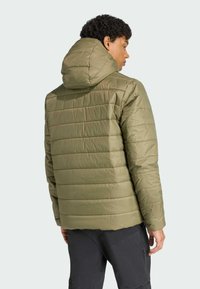 Personne portant une veste matelassée verte olive avec capuche, légèrement tournée loin de l'appareil photo, sur un fond clair.