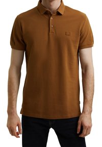 Polo shirt marron à manches courtes, avec patte de boutonnage à trois boutons et petit patch logo. Fabriqué en tissu texturé, associé à un jean bleu marine.