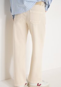 Vue arrière d'une personne portant un pantalon ample crème, une chemise rayée bleue et des baskets blanches avec un détail rouge, debout contre un mur blanc.