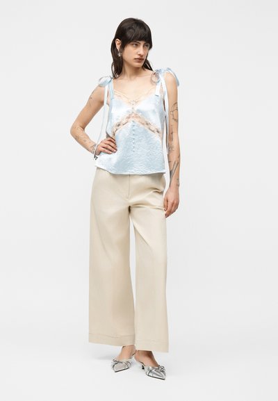 Γυναίκα με σκούρα μαλλιά, φοράει γαλάζιο σατέν καμιζόλ με δαντέλα, μπεζ παντελόνι φαρδύ στο κάτω μέρος και ασημί μυτερά τακούνια, στέκεται με το ένα χέρι στη μέση.