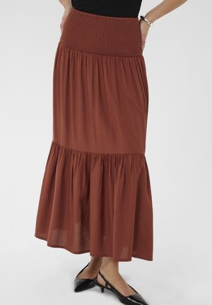 Femme portant une jupe longue plissée couleur rouille avec une taille smockée et des chaussures à talons bas noires à bride arrière, debout les mains sur les hanches.