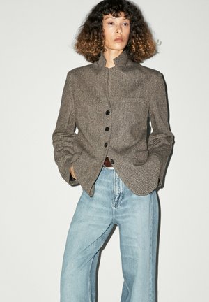 Femme aux cheveux bouclés portant un blazer marron boutonné et un jean bleu clair, debout les mains dans les poches devant un fond uni.