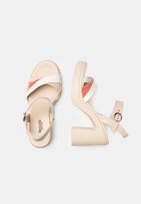 Sandali con tacco alto beige caratterizzati da un design a fascia incrociata con un accento lucido. Tacco a blocco grosso con una cinghia alla caviglia sicura e fibbia.