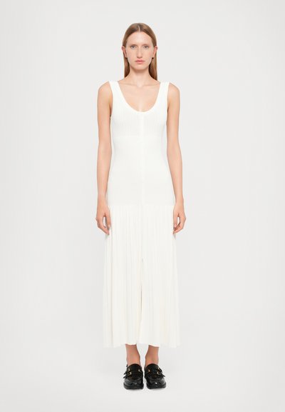 Robe en tricot côtelé blanche avec un décolleté en V profond, boutonnage devant et jupe plissée fluide. Associée à des chaussures noires à enfiler.