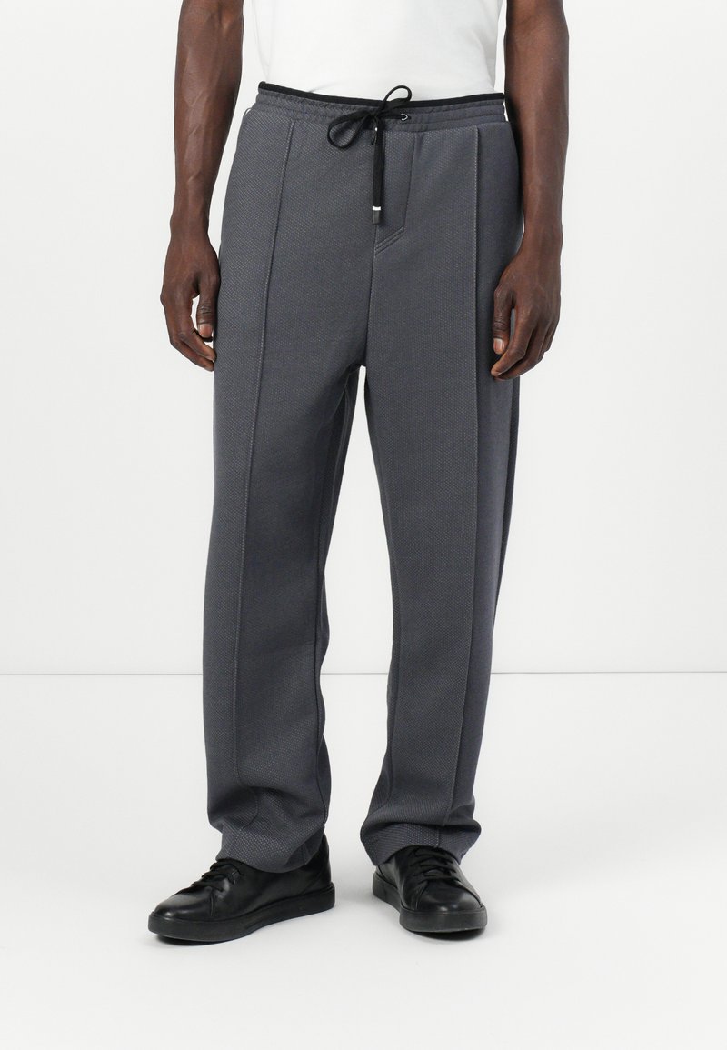 Pantaloni sport gri, cu un material texturat, talie elastică cu șnur de ajustare și două cusături frontale, asortați cu încălțăminte neagră.