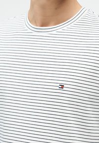 Κοντινό πλάνο ενός ατόμου που φοράει λευκό μπλουζάκι με λεπτές μαύρες οριζόντιες ρίγες και μικρό κόκκινο, άσπρο και μπλε λογότυπο στο στήθος.