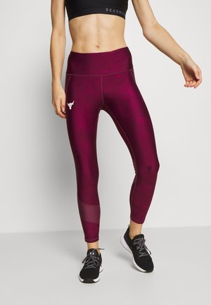 Femme portant un legging bordeaux taille haute avec un logo de taureau sur la cuisse, un soutien-gorge de sport noir et des chaussures de course noires sur un fond uni.
