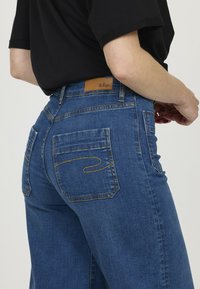 Jeans bleus taille haute en denim. Les caractéristiques incluent une étiquette en cuir à l'arrière, des coutures jaunes et deux poches arrière.