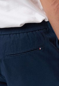 Közeli kép tengerészkék nadrágról, amelynek rugalmas derékrésze és kis trikolor címkéje van a hátsó zsebén, fehér inggel viselve.