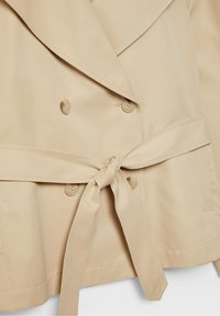Trench-coat beige à double boutonnage avec de larges revers, quatre boutons visibles et une ceinture nouée à la taille.
