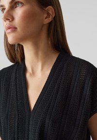 Top nero lavorato a maglia con scollatura a V, maniche corte e righe testurizzate. Il tessuto ha una lavorazione aperta, creando un design traspirante.