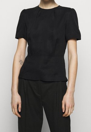 Personne portant un haut noir à manches courtes ajusté et un pantalon noir, avec un petit tatouage de soleil et de lune sur l'avant-bras gauche.