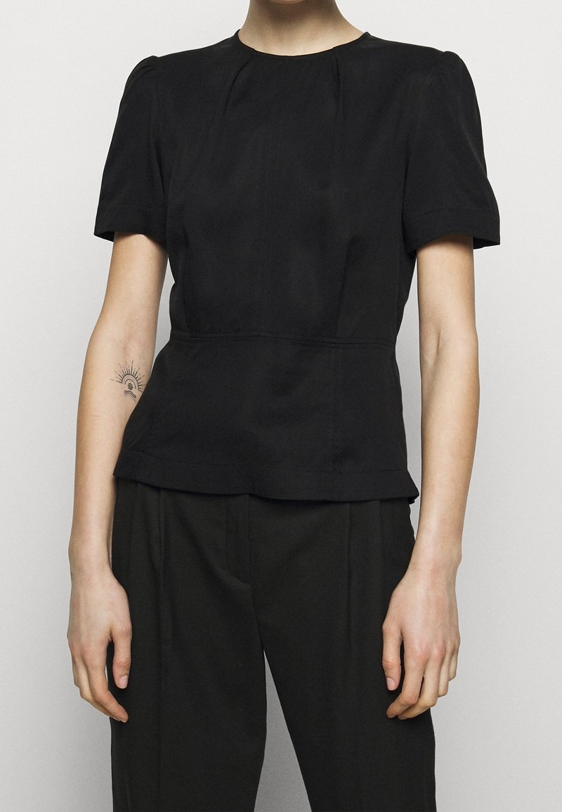 Osoba ubrana w dopasowany czarny krótki rękaw i czarne spodnie, z małym tatuażem słońca i księżyca na lewym przedramieniu.