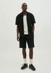 Férfi áll fekete rövid ujjú pólóban, hozzáillő fekete rövidnadrágban, fehér bordás trikóban, fehér zokniban és fekete sportcipőben egyszínű háttér előtt.