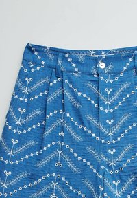 Tela azul con patrones florales y geométricos bordados en blanco, que incluye una pretina, trabillas para cinturón, botón y pliegues.