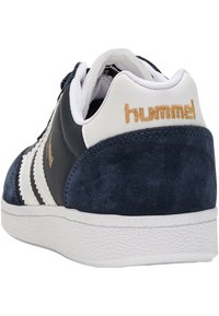 Mörkblå mocka och vit läder sneakers med guld "hummel"-logotyp på hälen, vita ränder och texturerad gummisula. Bakifrån syns snörningsdetaljer.