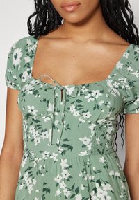 Zielona sukienka w kwiaty z krótkimi rękawami, kwadratowym dekoltem i marszczonym pasem. Materiał jest lekki, ozdobiony drobnymi białymi kwiatami i liśćmi.