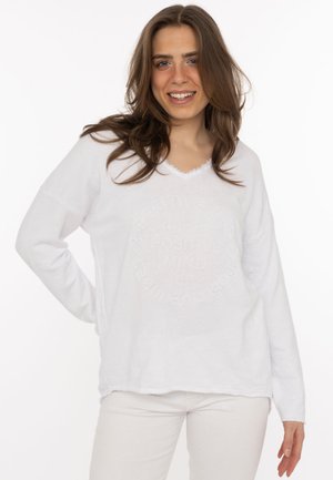 Young woman with long brown hair smiles wearing a white long-sleeve sweater and light-colored pants against a plain background.