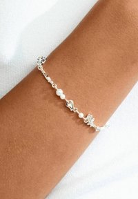 Silbernes Armband mit herzförmigen Gliedern und kleinen Perlen, auf einem braunen Arm platziert, vor einem strukturierten weißen Hintergrund.