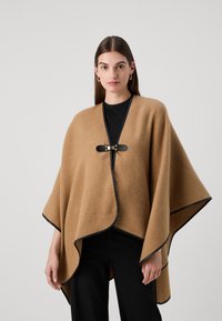 Lysbrun uldponcho med sort kant og en lukning foran. Har en flydende form og blød tekstur.