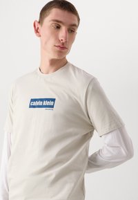 Beige bomull t-skjorte med korte ermer, med en blå rektangulær logo som har teksten "calvin klein" og "new york city" under.