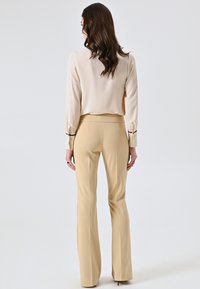 Beige Bluse mit langen Ärmeln und Manschetten, kombiniert mit hellbraunen, ausgestellten Hosen. Glatte Textur, minimalistisches Design, keine sichtbaren Muster oder Akzente.
